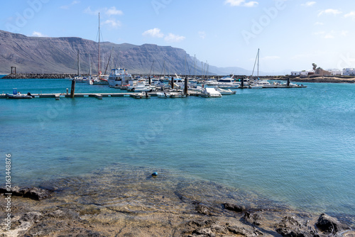 A scenic view of a marina on La Graciosa, Lanzarote, with boats docked and mountains in the background under a bright sky.