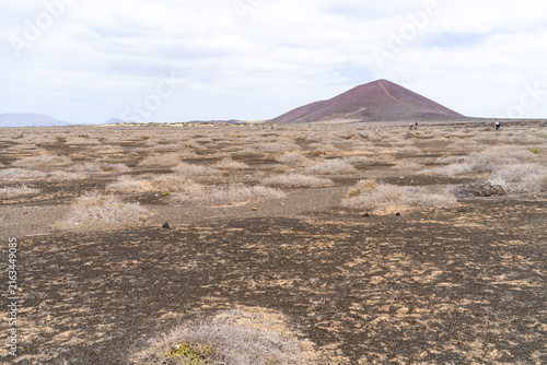 A view of a arid landscape on La Graciosa, with a volcanic mountain and people on the horizon. The sky is overcast with clouds.