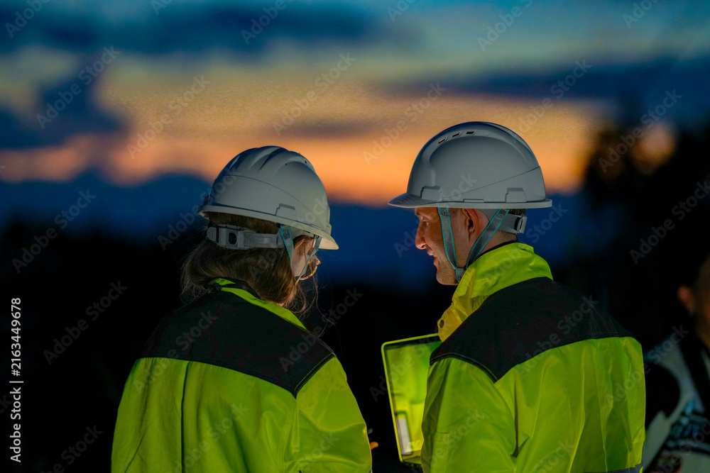 Fototapeta premium A technician and a female engineer read a tablet against the last orange sky in profile beneath the darkened rotor in vivid orange afterglow at a wind power plant in Southeast Asia.