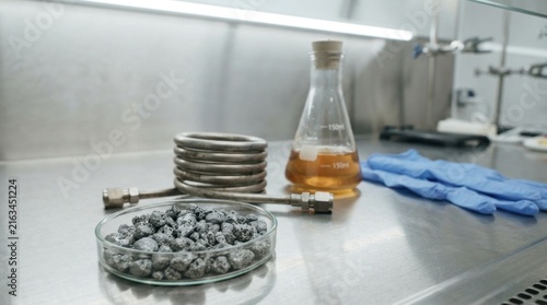 Heterogeneous catalysis concept with nickel catalyst granules, reactor tube, amber liquid flask, and blue nitrile gloves on a steel bench.