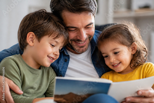 A father is reading a book to his two young children indoors, sharing a happy moment.