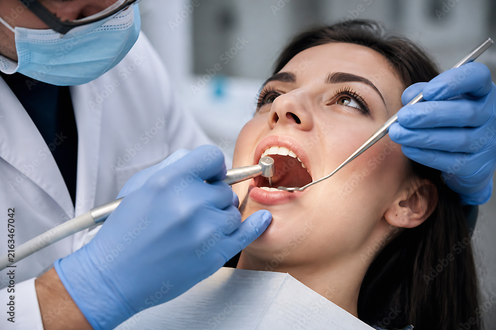 Naklejka premium A dentist in a white coat examines a female patient's mouth with a dental mirror and probe.
