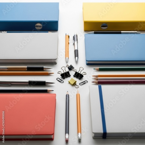 Organized flat lay of office supplies featuring blue and yellow ring binders, colorful notebooks, ballpoint pens, wooden pencils, and metal binder clips arranged on a white background.