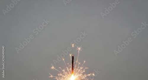 Single burning handheld sparkler firework with glowing tip and bright white sparks emitted from a dark stick, set against a neutral gray background with ample negative space.