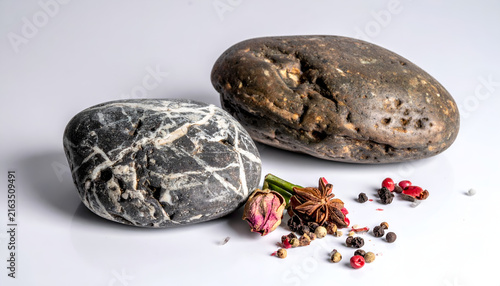 Close-up studio shot featuring two smooth stones, a sprig of dried flowers, star anise, and peppercorns on a bright, reflective white surface
