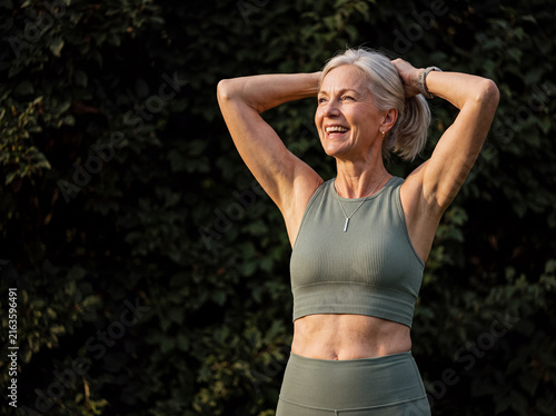 Happy senior woman in activewear smiling, with hands behind her head, enjoying outdoor exercise.