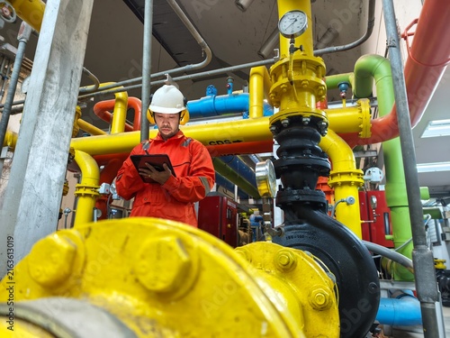 Cuadro en lienzo A low-angle shot of an industrial engineer in an orange jumpsuit, hardhat, and protective ear muffs holding a tablet while performing safety checks on a yellow gas pipeline system inside a facility