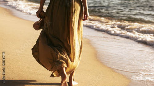 Young woman walking barefoot along a sandy beach shoreline at sunset, carryin...