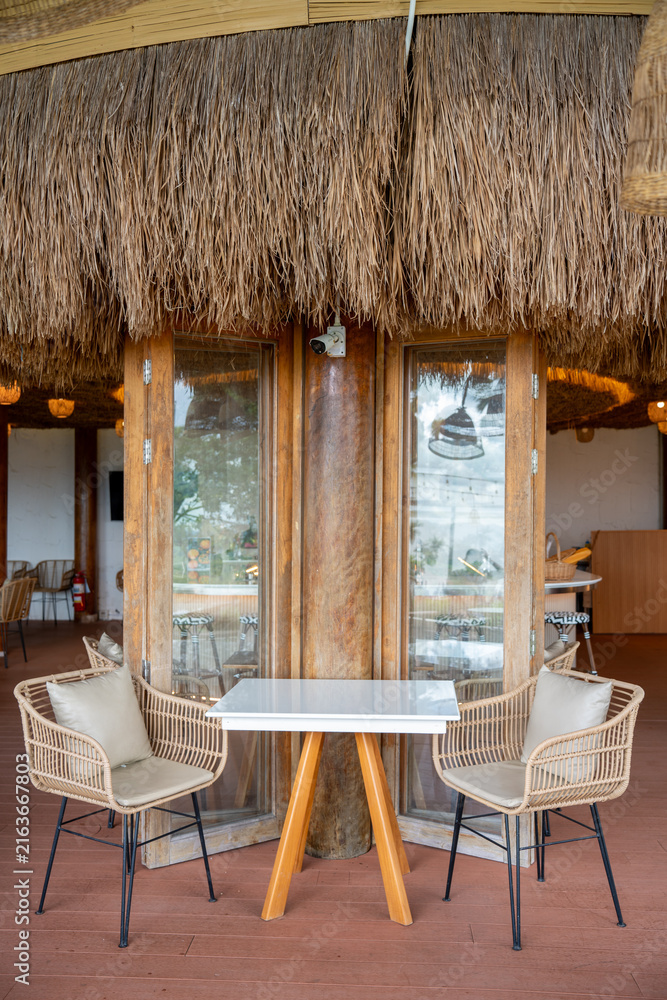 Fototapeta premium Empty cozy tropical cafe interior featuring a white table, woven rattan chairs with cushions, wooden flooring, and large wooden pillars against a white wall near glass folding doors.