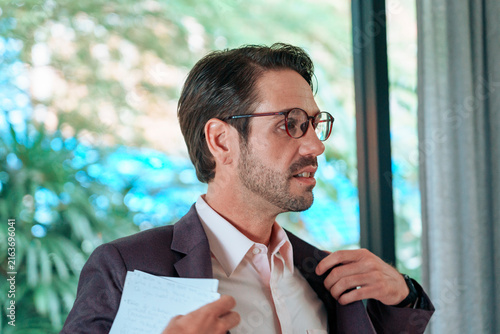 Foto A white man with glasses adjusting his clothes in a well-lit room with windows i