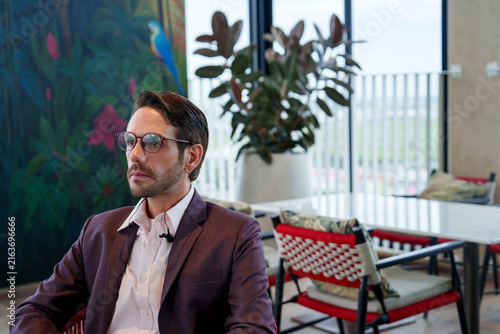 Cuadro en lienzo A white man with glasses in a stylish wine-colored suit sitting on a red chair i