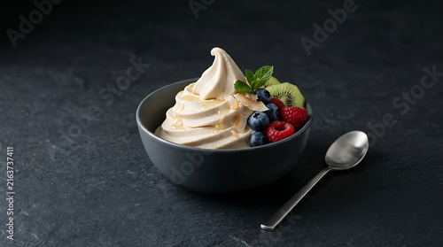 Creamy frozen yogurt swirl topped with fresh blueberries raspberries kiwi and mint leaves drizzled with honey in a dark grey bowl on a slate background with a silver spoon nearby