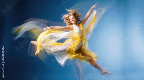 Happy young woman dancing in a flowing yellow dress with long fabric motion blur isolated on a blue studio background
