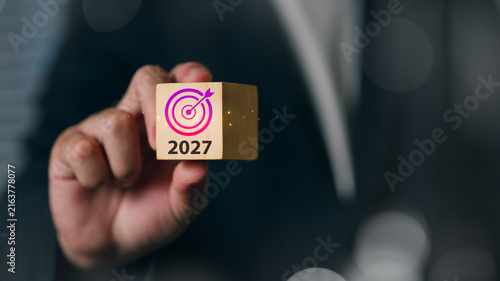 A professional businessman presenting a wooden cube with a target symbol and the year 2027, symbolizing future goals, planning, and strategic success.