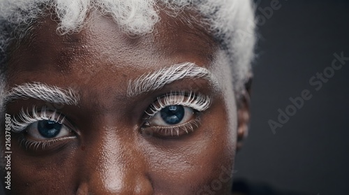 Detailed close up portrait of a black person featuring unique white eyelashes, white eyebrows, and curly white hair. The striking blue eyes create a powerful contrast against the rich dark skin ton...