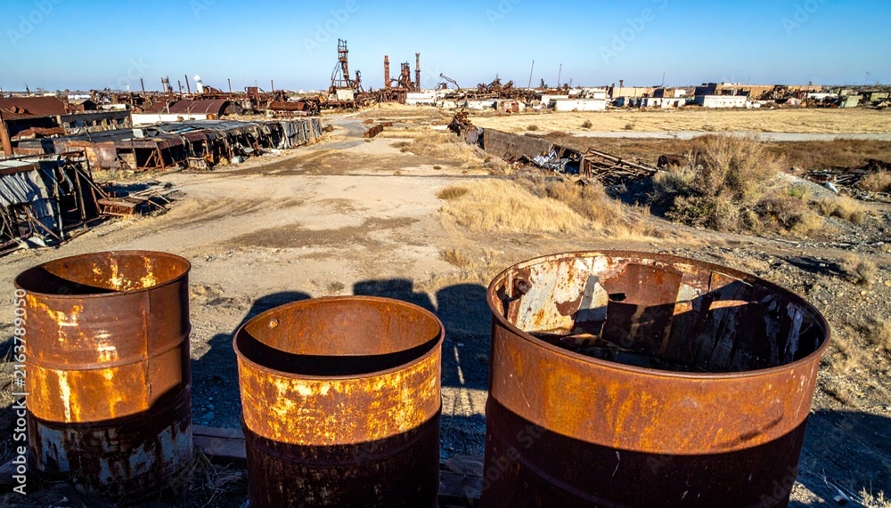 Naklejka premium Abandoned industrial wasteland with rusty barrels in the foreground and derelict factory structures in the background under a clear blue sky.