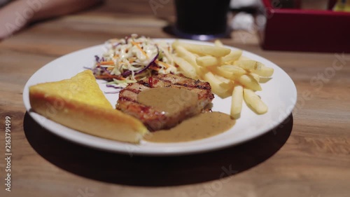 Grilled pork steak with black pepper gravy served with crispy french fries, coleslaw, and toasted buttered bread on a white plate. Close-up of a delicious restaurant meal on a wooden table.