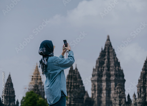 Bild auf Leinwand Muslim Asian woman traveler capture a picture of vacation, travel and tourism with smartphone standing on the tourism destination in Prambanan temple, Yogyakarta, Indonesia