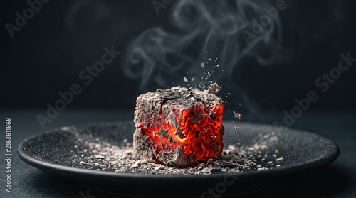 Glowing Embers: A single cube of burning charcoal smolders on a dark plate, releasing wisps of smoke into the air. The intense red heat and ash create a dramatic, evocative image.