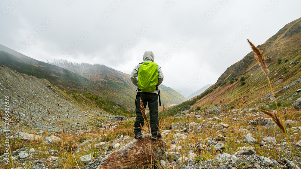 Obraz premium Hiker with green backpack standing on rock looking at autumn mountain valley near Shkhara glacier in Georgia.