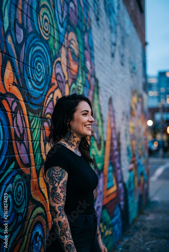 Young woman with tattoo smiling at colorful street graffiti wall wearing black
