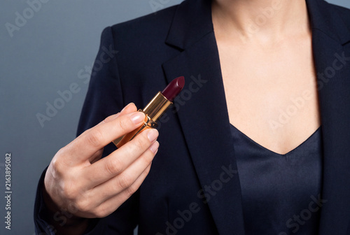 A business woman in a dark navy blazer holding open dark burgundy lipstick tube against grey studio background. Professional beauty and makeup product concept