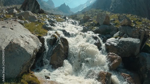 Rushing white water flows through rocky alpine valley under clear blue sky. Sunlight highlights mist spray. Composition leads eye through rugged terrain with distant peaks