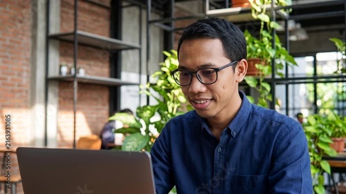 Smiling Indonesian business man looking at laptop using computer working in office. Busy professional businessman employee or company executive searching online solutions at workplace.