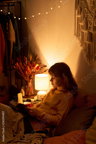 Vertical image of Caucasian young adult woman reading book in bed under warm lamp light, promoting mental wellbeing, self care, and cozy evening relaxation concepts
