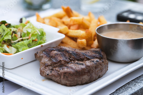 Canvastavla Grilled beef steak served with french fries, fresh side salad and pepper sauce on a white plate in an outdoor restaurant