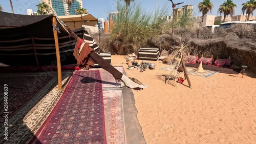 Wide shot of a Bedouin tent with rugs, a fire pit, and personal belongings in Dubai, with skyscrapers visible in the background during daytime.