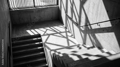 Sunlight streams into a concrete staircase, casting dramatic shadows across the interior space. The staircase leads upwards, disappearing into the darkness.
