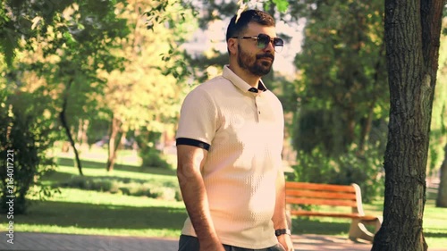Various video clips of a bearded man wearing sunglasses and a polo shirt walking in a green park. He strolls along a paved path and adjusts his sleeve under natural daylight among trees.