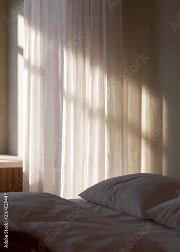 Bedroom with a bed in the foreground. the bed has white sheets and two white pillows. the window is covered with sheer white curtains that are open, creating a soft and airy atmosphere.