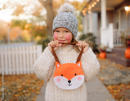 A cute young girl in a cozy sweater and beanie holding a fox-shaped trick-or-treat bag.