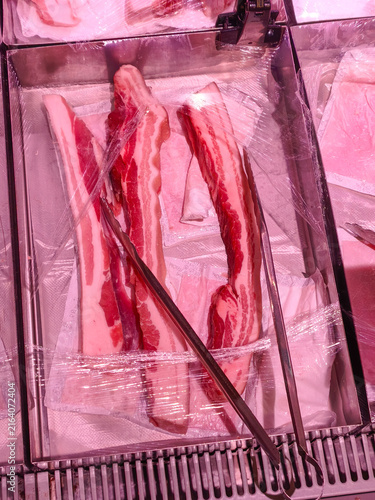 Fresh Pork Belly Strips in Display Case. High-angle shot of raw, marbled pork belly strips resting in a butcher counter tray under pink lighting, ready for grilling.