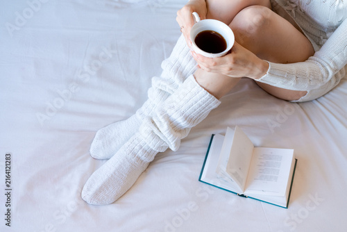 Fotografie Young caucasian woman holding a porcelain cup of black coffee beside an open book on a bed, fits morning ritual, cafe and slow living content