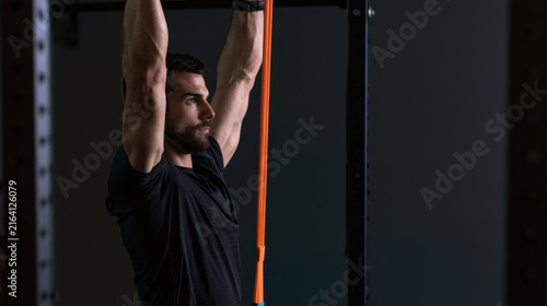 A man stands in a gym and pulls down a resistance band attached to a bar. He is focused on his workout. Light from the side highlights his effort