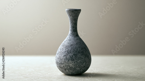 A stone vase stands on a flat surface with a plain background. The surface is light and the vase has a smooth, rounded shape. The scene is simple and minimal