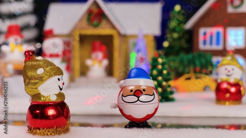 A quirky Santa Claus jumps up to a snowman against a blurred background of a Christmas village