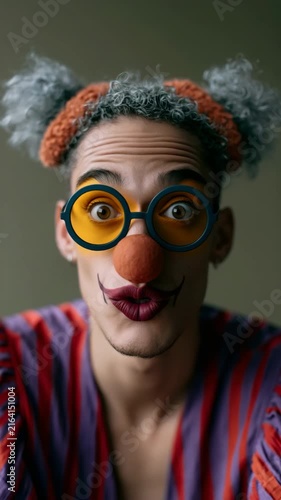 Vertical portrait of a laughing person in striped clown outfit with orange nose and oversized round yellow glasses for poster background and playful editorial use