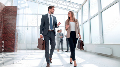 Happy young businessman and businesswoman walking and talking together in a bright modern glass office corridor with briefcases.