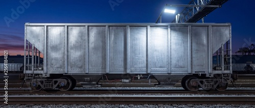 Metal hopper railcar parked on industrial tracks at night under bright overhead security lighting with other train wagons in background