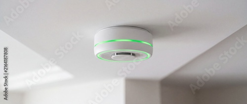 Modern white smoke detector with glowing green led status ring mounted on clean ceiling provides reliable fire safety monitoring in homes