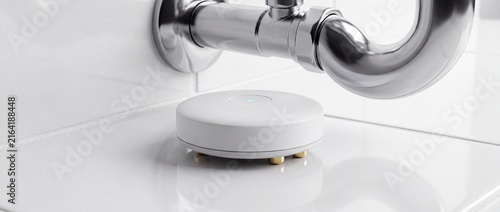White circular smart water leak detector sensor sits on shiny floor tile under chrome metal bathroom sink pipes protecting home interiors