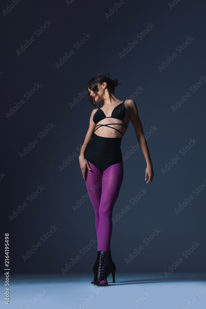 Fototapeta premium Full length image of a sexy, beautiful dancer dressed in a sensual outfit with purple tights, moving in dance poses in the studio, against a dark background.