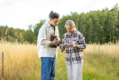 Agricultural analysts stand together studying data on tablet with focused expressions in wheat field. Concept of thorough agricultural analysis and collaborative teamwork between farming colleagues.