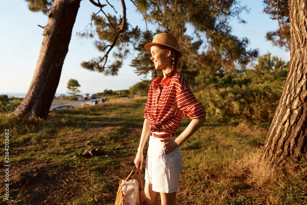 Naklejka premium Young woman enjoying lifestyle outdoor nature portrait. She wears straw hat and striped shirt standing in park forest at sunset. Summer mood concept for travel and vacation.
