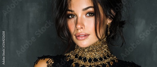 A portrait of a woman with dark hair wearing an elaborate gold necklace against a dark background.