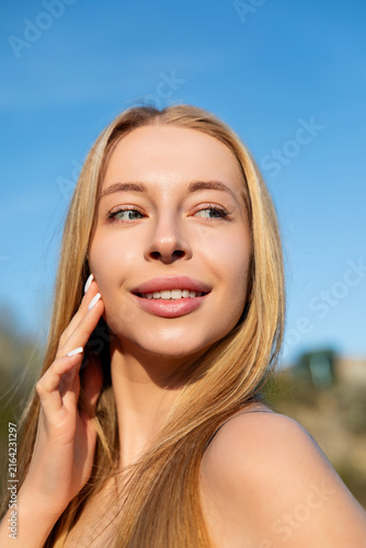 Beauty woman smiling and touching her smooth cheek while looking into the distance. Attractive woman portrait reveals radiant skin, natural beauty and skincare concept under blue sky.